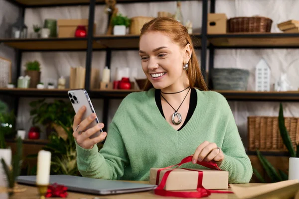 Una giovane donna accogliente sorride mentre apre un regalo e si gode un momento sul suo telefono a casa.. — Foto stock