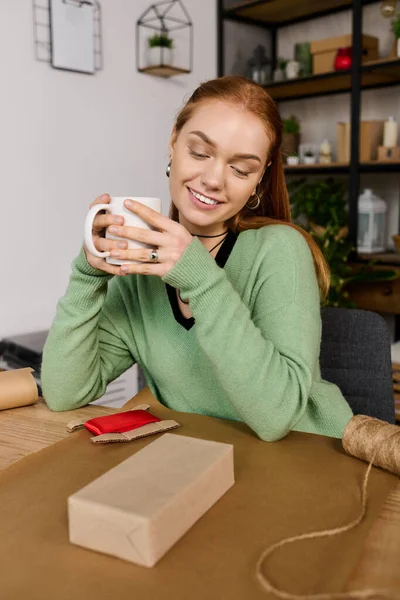 Young woman enjoys sipping a warm drink while sitting at her cozy home workspace. — Stock Photo