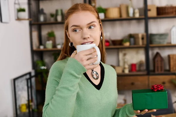 Eine junge Frau entspannt sich zu Hause, nippt an einem warmen Getränk und hält ein schön verpacktes Geschenk in der Hand.. — Stockfoto