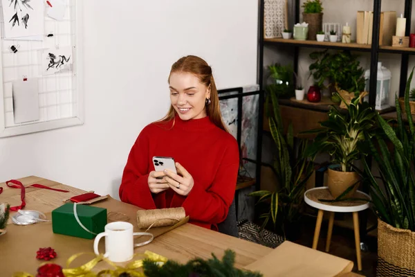 Молода жінка, одягнена в червоний светр посміхається, використовуючи свій смартфон в затишній домашній обстановці. — стокове фото