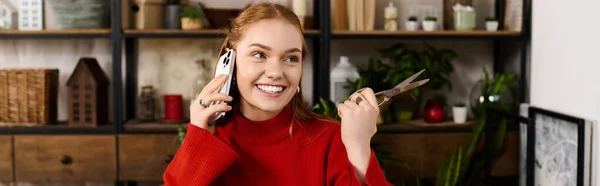 Joyful young woman at home, chatting on the phone while holding scissors, surrounded by cozy decor. — Stock Photo