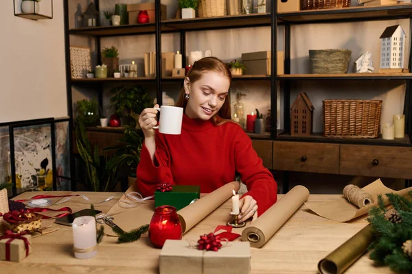 Насолоджуючись затишним днем, молода жінка обнімає подарунки, смакуючи свій теплий напій. — стокове фото