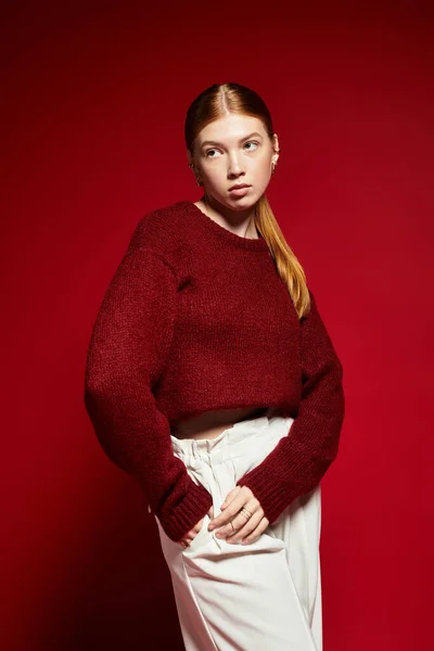 Una joven con el pelo rojo posa con confianza en un acogedor suéter de invierno sobre un llamativo fondo rojo. - foto de stock