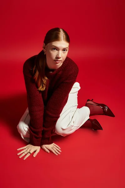 Une jeune femme pose ludique dans un pull d'hiver sur un fond rouge audacieux, mettant en valeur son style. — Photo de stock