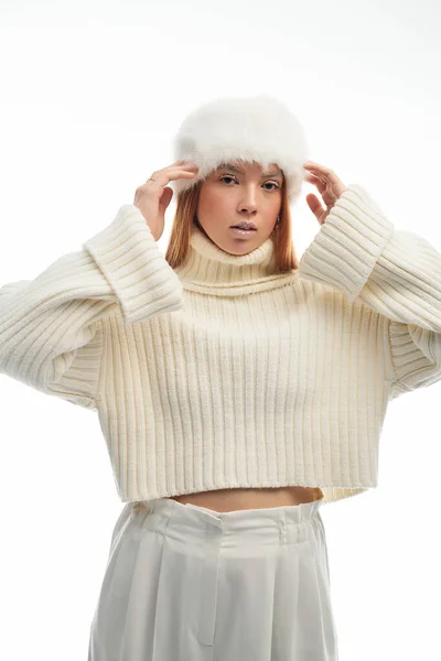 Young redhead woman poses confidently in a cozy knit sweater and fur hat against a white background. — Stock Photo