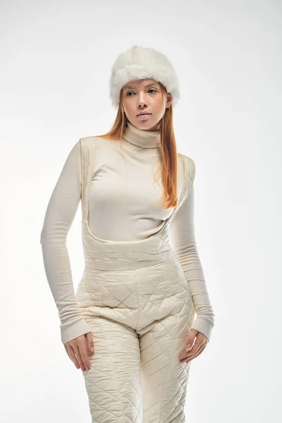 Une jeune rousse pose élégamment dans un pull confortable et un chapeau de fourrure sur un fond blanc vif. — Photo de stock