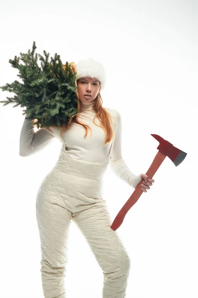 Une jeune rousse pose dans un pull confortable et un chapeau de fourrure, tenant un arbre de Noël festif. — Photo de stock