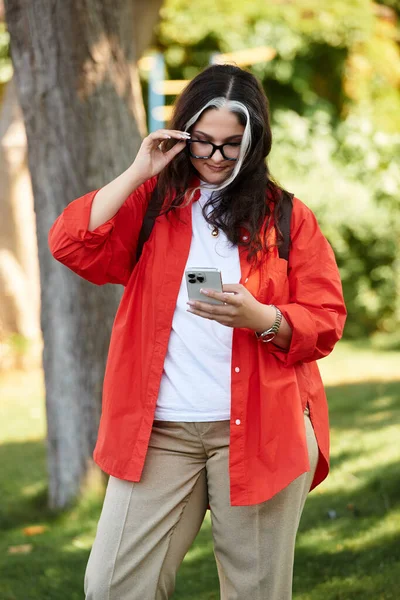 A woman with a unique white forelock is outside, happily using her smartphone in nature. — Stock Photo