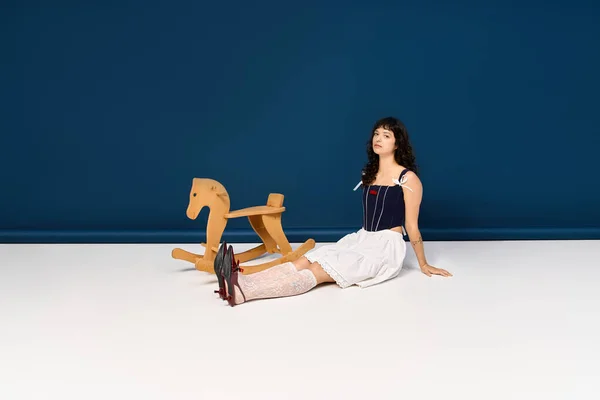 Eine junge, schöne Frau sitzt elegant neben einem hölzernen Schaukelpferd auf einem sauberen weißen Boden.. — Stockfoto