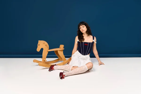 Стильная молодая женщина изящно сидит рядом с деревянной качающейся лошадью в современном пространстве. — стоковое фото