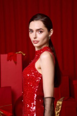 Güzel genç bir kadın zarif bir şekilde parıldayan kırmızı elbiseyle poz veriyor. Etrafı şenlikli dekorasyonlarla çevrili..