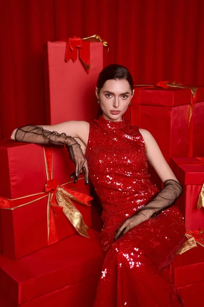 Göz kamaştırıcı kırmızı elbiseli genç bir kadın renkli hediye kutularının yanında oturur ve eğlencenin tadını çıkarır..