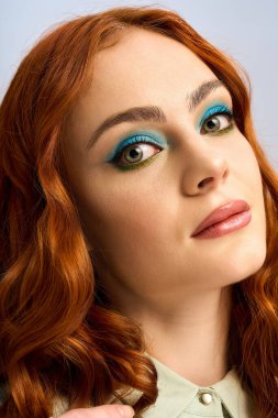 Genç bir kızıl, kendine güvenen bir şekilde stüdyoda poz veriyor, göze çarpan cesur göz makyajı yapıyor..