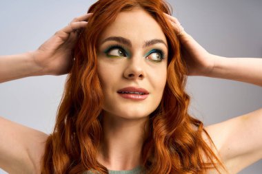 Genç kızıl saçlı, göz kamaştırıcı makyaj sanatını vurgulayarak stüdyoda şakacı bir poz veriyor..
