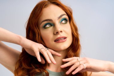 Kızıl saçlı genç kadın zarif poz veriyor, canlı makyajını ve neşeli ifadesini vurguluyor..