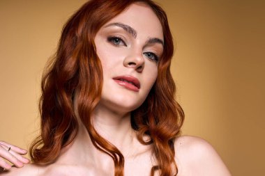Kızıl saçlı, kendine güvenen genç bir kadın stüdyoda poz veriyor, yumuşak makyajını sergiliyor..