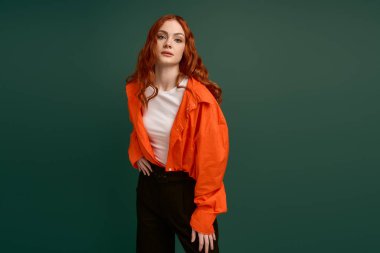 Kızıl saçlı genç bir kadın turuncu gömlekli, kendine güvenen bir poz veriyor, sonbahar modası için mükemmel..