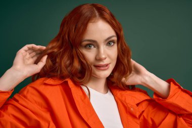 Turuncu tişörtlü genç bir kızıl yeşil fonda kendine güvenen bir poz veriyor..