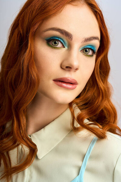 Молодая женщина с яркими рыжими волосами и ярким макияжем глаз уверенно позирует в студии.