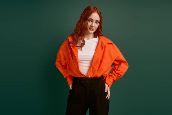 Рыжая женщина носит оранжевую рубашку на пышном зеленом фоне, воплощая осенний стиль.