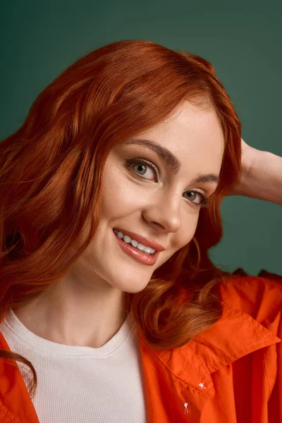Kızıl saçlı bir kadın turuncu bir gömlekle yeşil bir arka plana gülümsüyor, sonbahar tarzını sergiliyor..