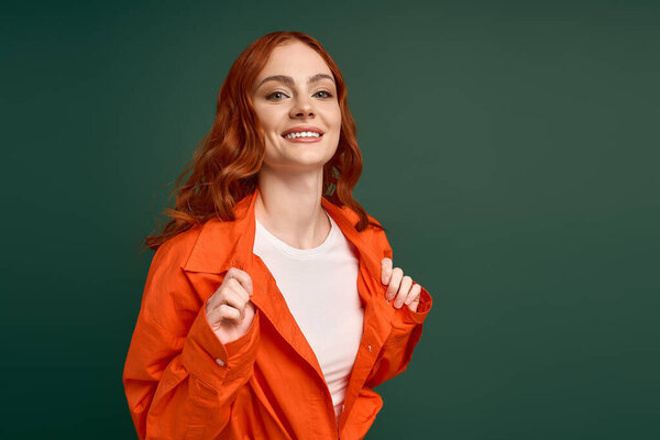 A young redhead woman smiles while wearing an orange shirt against a green background.