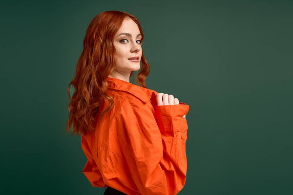 A young woman with red hair poses confidently in an orange shirt against a green background.