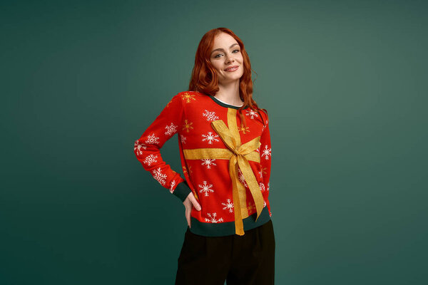 Радостная рыжая девушка демонстрирует свой рождественский свитер на ярком зеленом фоне..