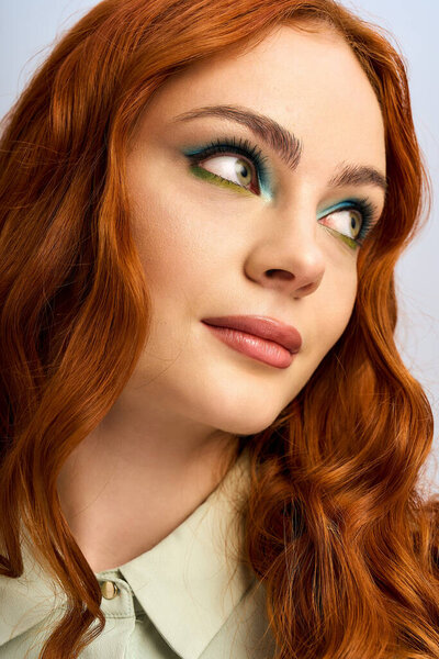 Молодая женщина с яркими рыжими волосами демонстрирует яркий макияж глаз на сером фоне.