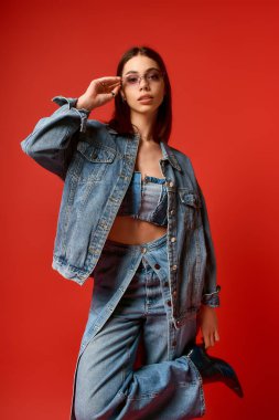 Genç bir kadın, parlak kırmızı bir arka planda kot giyip güvenle poz veriyor..