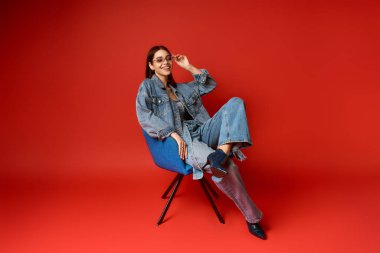 Modayı takip eden kot elbiseli genç bir kadın, mavi bir sandalyede, parlak kırmızı bir arka planda dinleniyor..