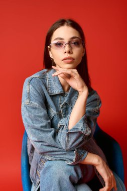 Kot ceketli ve kot pantolonlu genç bir kadın kırmızı arka planlı bir stüdyoda zarif pozlar veriyor..