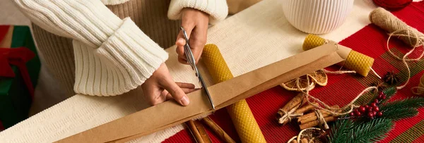 Hands skillfully cut wrapping paper to prepare Christmas gifts surrounded by festive decor. — Stock Photo