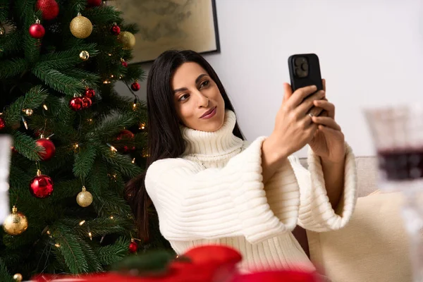 Una mujer disfruta tomando una selfie rodeada de decoraciones navideñas y un árbol bellamente adornado. - foto de stock