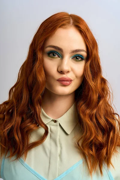 Une femme rousse pose avec confiance, affichant un maquillage audacieux des yeux et des boucles vibrantes dans un studio. — Photo de stock