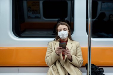 Koruyucu tek kullanımlık maske takan güzel bir kadın sosyal mesafeyi koruyor ve taşınırken yalnız başına oturarak akıllı telefon kullanıyor. Yeni normal yaşam tarzı kavramı..