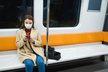 Koruyucu tek kullanımlık maske takan güzel bir kadın sosyal mesafeyi koruyor ve taşınırken yalnız başına oturarak akıllı telefon kullanıyor. Yeni normal yaşam tarzı kavramı..