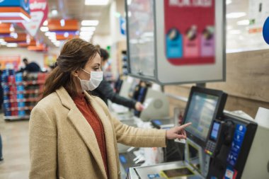 Tek kullanımlık koruyucu maske takan güzel bir kadın alışveriş merkezinde alışveriş için para ödüyor. Salgın sırasında yeni normal konsept, covid19.
