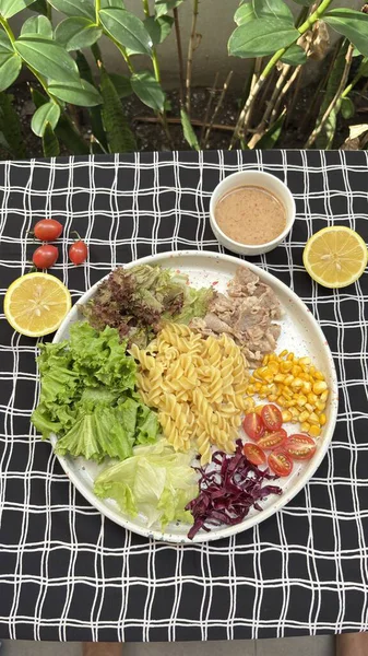 A colorful fresh vegetable salad with pasta cherry tomatoes and lemon dressing served on a white plate on black and white checkered tablecloth in natural lighting conveying healthy and vibrant cuisine for food and recipe marketing.