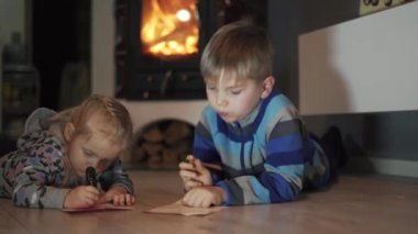İki Küçük Mutlu Çocuk Kardeş ve Kız Kardeş Odada Yerde Yatıyor Yazıyor ve Çiziyor. Arka planda yanan şömineli rahat atmosfer. Mutlu Aile kavramı. Doli Vuruşu
