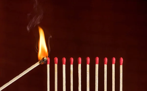 Lit match next to a row of unlit matches. Red phosphorus matches on dark red background. Concept of ignition or initiation