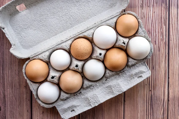 A cardboard egg box with ten brown and white eggs on a wooden table. Mixed eggs in a box - Top View