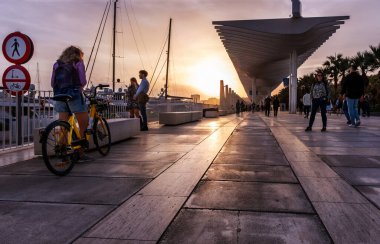 Malaga, İspanya - 15 Kasım 2024: Malaga Marina Promenade, Endülüs, İspanya gün batımında. Paseo del Muelle Dos Gezinti Alanı boyunca yürüyen insanlar, beyaz dalgaları andıran bir yapı altında limana doğru ilerliyorlar.
