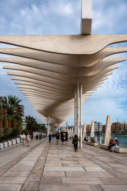 Malaga, İspanya - 15 Kasım 2024: Malaga Marina Promenade, Endülüs, İspanya gün batımında. Paseo del Muelle Dos Gezinti Alanı boyunca yürüyen insanlar, beyaz dalgaları andıran bir yapı altında limana doğru ilerliyorlar.