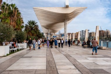 Malaga, İspanya - 15 Kasım 2024: Malaga Marina Promenade, Endülüs, İspanya gün batımında. Paseo del Muelle Dos Gezinti Alanı boyunca yürüyen insanlar, liman boyunca uzanan beyaz dalgalara benzeyen bir yapı altında