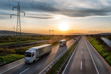 Güzel bir gün batımında, otoyolda kırmızı konteynırlarla beyaz kamyonlar konvoyu, ilkbaharda kargo taşıma konsepti - nakliye hizmeti