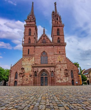 Basel 'deki tarihi Gotik katedralde ikiz kuleler, taş cephe, kaldırım taşı meydan ve dramatik gökyüzü.
