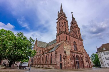 Basel 'deki tarihi Gotik katedralde ikiz kuleler, taş cephe, kaldırım taşı meydan ve dramatik gökyüzü.