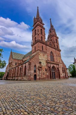 Basel 'deki tarihi Gotik katedralde ikiz kuleler, taş cephe, kaldırım taşı meydan ve dramatik gökyüzü.