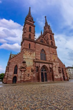 Basel 'deki tarihi Gotik katedralde ikiz kuleler, taş cephe, kaldırım taşı meydan ve dramatik gökyüzü.